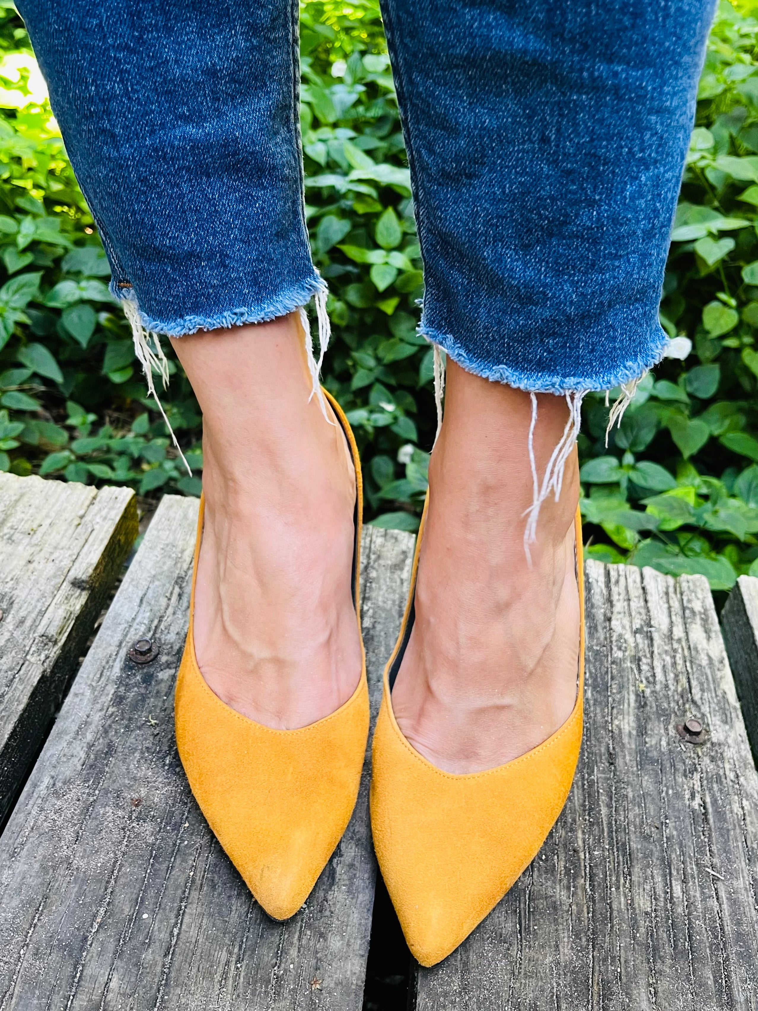Yellow pointed-toe suede shoes worn with blue jeans on a wooden surface with greenery in the background